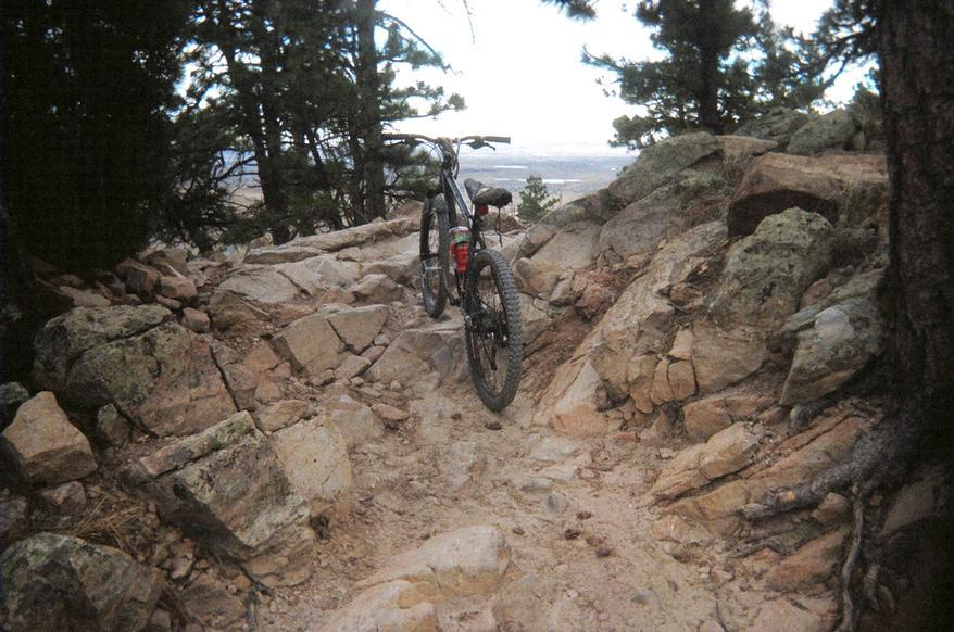 A mountain bike standing on a rugged, rocky trail surrounded by trees, with a scenic view in the background. The terrain features loose rocks and gravel, illustrating a challenging path for biking. Red Rocks / Dakota Ridge mountain bike trail.