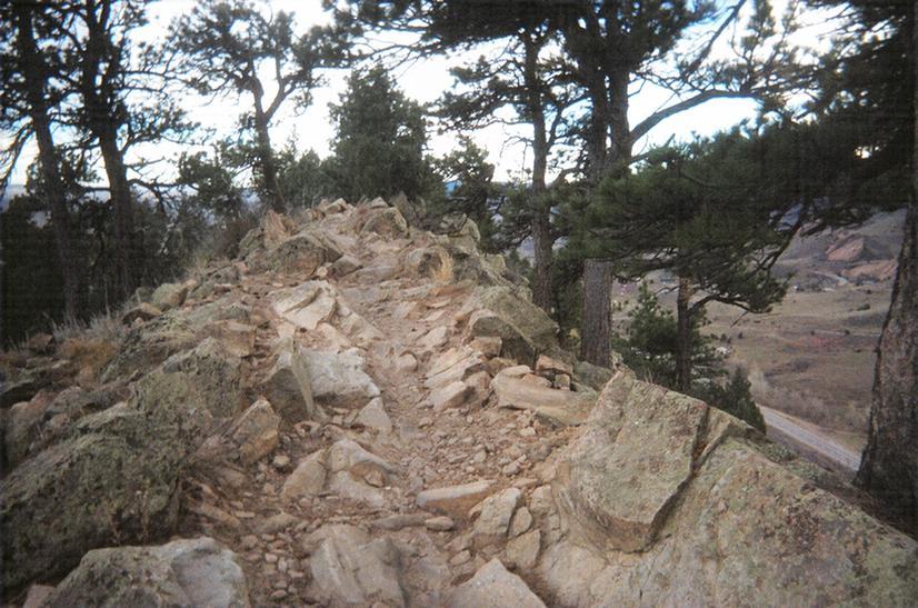 A rocky hiking trail bordered by trees, leading along a ridge with a view of a valley below. The terrain is uneven with various sizes of stones and pebbles. Red Rocks / Dakota Ridge mountain bike trail.