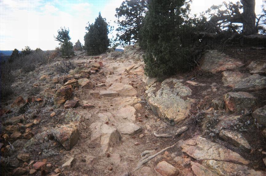 "Rocky hiking path winding through rugged terrain, bordered by sparse vegetation and trees, under a cloudy sky." Red Rocks / Dakota Ridge mountain bike trail.