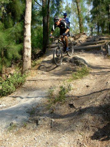 Mountain biker navigating a rocky trail through a forested area, with another cyclist in the background. The rider is captured mid-jump, showcasing an action-packed moment in nature. Markham Park mountain bike trail.