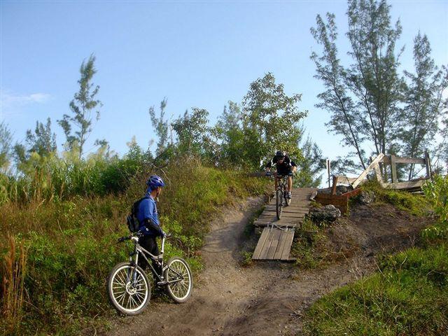 Two mountain bikers navigate a dirt trail with a wooden plank bridge in a natural outdoor setting. One cyclist is ascending the bridge while the other is positioned at the bottom of the trail, wearing a blue helmet and preparing to ride. Surrounding vegetation includes tall grasses and trees under a clear blue sky. Markham Park mountain bike trail.