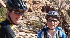Two cyclists wearing helmets and sunglasses, posing for a photo outdoors on rocky terrain. The background features natural rock formations and sparse vegetation. Klondike Bluffs mountain bike trail.