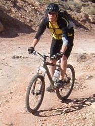 A person riding a mountain bike along a rocky trail, wearing a helmet and athletic gear in yellow and black. The terrain is rough and dusty, with natural rock formations in the background. Klondike Bluffs mountain bike trail.
