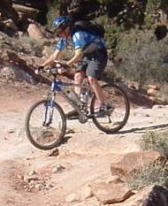 A person riding a mountain bike on a rocky trail surrounded by vegetation. The rider is wearing a blue shirt, shorts, and a helmet, navigating a challenging terrain. Klondike Bluffs mountain bike trail.
