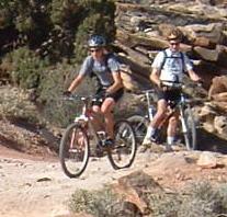 Two mountain bikers riding on a rocky dirt trail surrounded by shrubs and boulders. One rider is in the foreground, actively biking, while the other follows behind. Both are wearing helmets and cycling gear. Klondike Bluffs mountain bike trail.