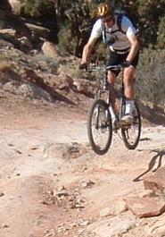 A mountain biker in mid-air, jumping off a rocky terrain on a mountain trail, wearing a helmet and cycling gear. Klondike Bluffs mountain bike trail.