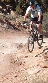 A mountain biker in a helmet jumps over rocky terrain on a trail surrounded by sparse vegetation. Klondike Bluffs mountain bike trail.