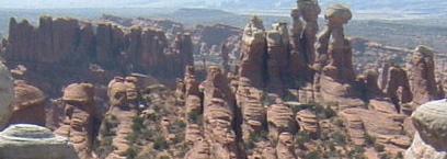 A panoramic view of rugged, red rock formations in a desert landscape, featuring tall, slender spires and unique geological shapes against a distant backdrop of rolling hills and valleys. Klondike Bluffs mountain bike trail.