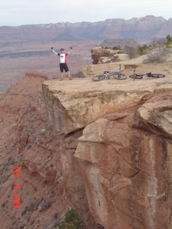 A person stands triumphantly on the edge of a rocky cliff, with arms raised in celebration. Two mountain bikes are parked nearby on the plateau, and the expansive desert landscape stretches out behind them, showcasing layers of red rock formations and distant mountains under a cloudy sky. Gooseberry Mesa mountain bike trail.