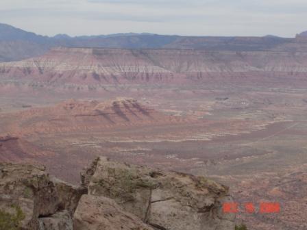 A panoramic view of a rugged desert landscape featuring layered rock formations in shades of red and brown, with distant mountains under a cloudy sky. Gooseberry Mesa mountain bike trail.
