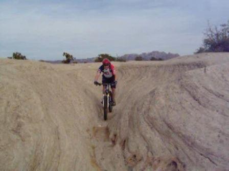 A mountain biker navigating a narrow, sandy trail in a desert landscape, with rocky hills in the background and sparse vegetation nearby. Gooseberry Mesa mountain bike trail.