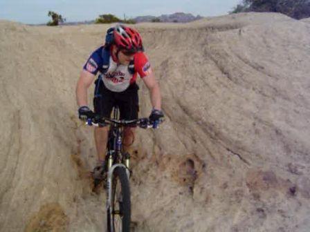 A mountain biker navigating a rocky terrain, focusing on maintaining balance while riding. The rider is wearing a red helmet and a short-sleeve cycling jersey, and is surrounded by natural landscape features. Gooseberry Mesa mountain bike trail.