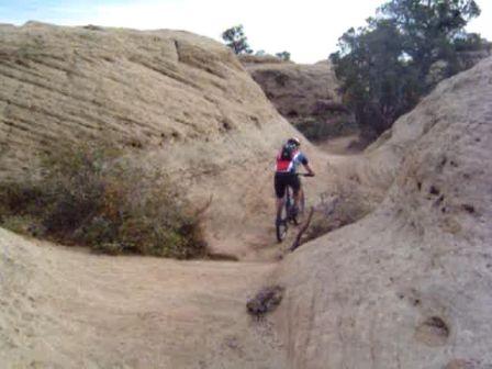 A person riding a mountain bike through a rugged, rocky trail surrounded by steep, sandy terrain and sparse vegetation. The landscape features natural formations and a clear sky in the background. Gooseberry Mesa mountain bike trail.