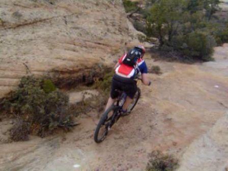 A cyclist riding down a rocky, uneven trail surrounded by vegetation and rugged terrain. The rider is wearing a backpack and is seen from the back, navigating the challenging landscape. Gooseberry Mesa mountain bike trail.
