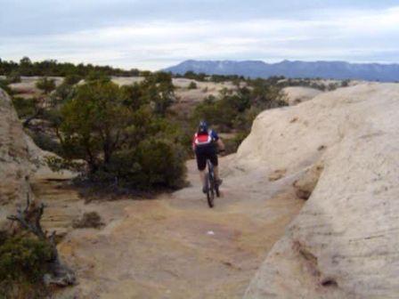 A person riding a mountain bike along a narrow trail surrounded by rocky terrain and sparse vegetation, with distant mountains visible under a cloudy sky. Gooseberry Mesa mountain bike trail.