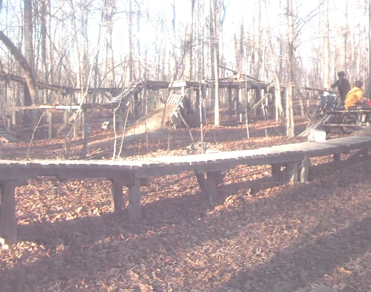 A wooded area with a wooden path leading through fallen leaves, featuring a bike parked beside a picnic table. In the background, there are wooden structures and ramps, suggesting a recreational or outdoor activity space. The scene is illuminated by soft sunlight filtering through the trees. Pee Wee's Mountain Bike Park mountain bike trail.