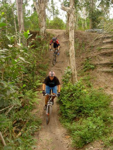 Two mountain bikers navigating a dirt trail through a lush, green forest. One rider is descending a slope while the other is positioned further up the trail. The surrounding area features tall trees and dense vegetation, creating a natural outdoor setting. Markham Park mountain bike trail.