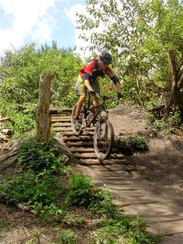 A cyclist navigating a wooden bridge on a mountain bike trail, surrounded by lush greenery and trees. The rider is wearing a helmet and athletic gear, and the terrain is earthy with patches of grass and foliage. Markham Park mountain bike trail.