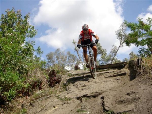 A mountain biker riding down a dirt trail surrounded by greenery, with a partly cloudy sky in the background. The cyclist is wearing a red and white jersey and a helmet, focusing on navigating the challenging terrain. Markham Park mountain bike trail.