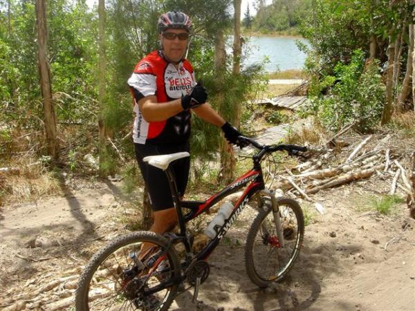 A cyclist wearing a red and white jersey and a helmet gives a thumbs-up while standing next to a mountain bike, surrounded by trees and a river in the background. Markham Park mountain bike trail.