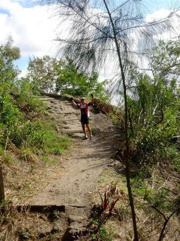 A person standing triumphantly on a trail surrounded by lush greenery, with a clear sky above. The individual is wearing a sports outfit and appears to be celebrating their hike. Steps are visible leading up the trail. Markham Park mountain bike trail.