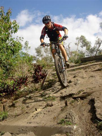 A mountain biker navigating a steep, dirt trail surrounded by greenery and trees under a partly cloudy sky. Markham Park mountain bike trail.