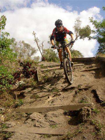 A mountain biker descending a rugged, dirt trail surrounded by greenery, wearing a helmet and sports attire, with a mixture of dirt and rocks on the path. Markham Park mountain bike trail.