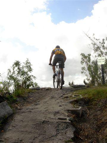 A person riding a mountain bike uphill on a dirt trail, surrounded by greenery, with a caution sign visible in the background under a partly cloudy sky. Markham Park mountain bike trail.