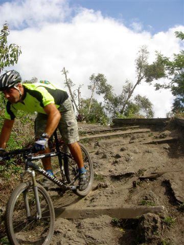A mountain biker navigating a rocky, uneven trail with trees and a blue sky in the background. Markham Park mountain bike trail.