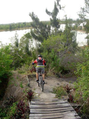 A person riding a mountain bike down a narrow dirt path surrounded by greenery, with a river visible in the background. The rider is wearing a red and black cycling outfit and is navigating a wooden bridge. Markham Park mountain bike trail.