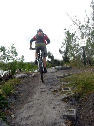 A person riding a mountain bike on a dirt trail surrounded by greenery, heading uphill. A caution sign is visible in the background, and the trail is lined with rocks. The sky is partly cloudy. Markham Park mountain bike trail.
