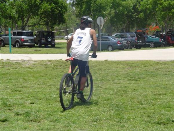 A person wearing a helmet and a white tank top with the number 7 is riding a bicycle on a grassy area, with parked cars and trees in the background. Markham Park mountain bike trail.