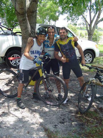 Three young men posing with mountain bikes next to a tree. Two of them are wearing cycling attire, while the third is in a basketball jersey. A white SUV is parked in the background. The setting appears to be outdoors, possibly at a biking or recreational area. Markham Park mountain bike trail.