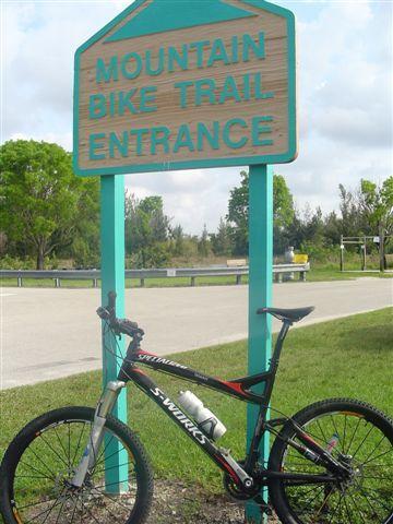 A mountain bike leaning against a sign that reads "Mountain Bike Trail Entrance," with a grassy area and trees in the background. The pathway leads into the trail, indicating an outdoor recreational area. Markham Park mountain bike trail.