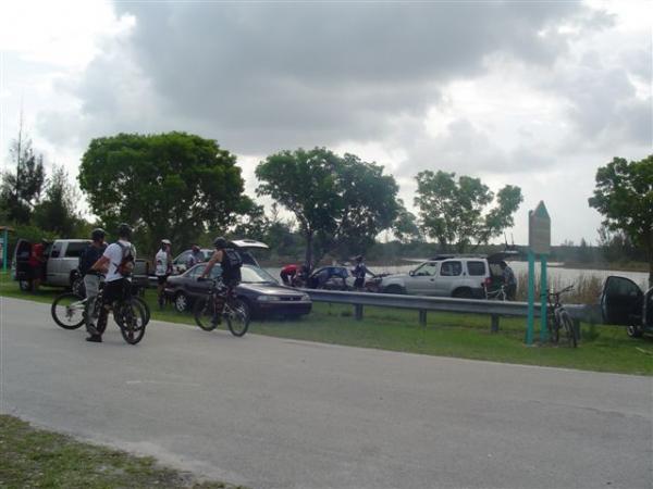 A group of cyclists in casual attire stands by their bicycles near a parking area. Several vehicles are parked alongside a road, with a backdrop of trees and a body of water in the distance. The sky is partly cloudy, creating a serene outdoor atmosphere. Markham Park mountain bike trail.