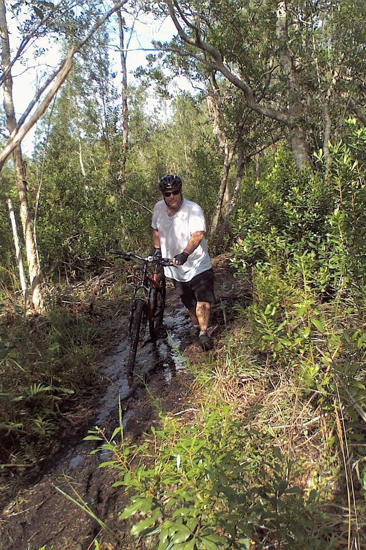 A cyclist standing in a muddy trail surrounded by trees, pushing a mountain bike. The cyclist is wearing a helmet, sunglasses, and gloves, and is dressed in a white t-shirt and shorts. Sunlight filters through the foliage, illuminating the damp, grassy ground. Markham Park mountain bike trail.
