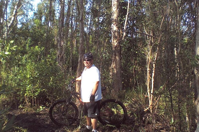 A person wearing sunglasses and a white T-shirt stands next to a mountain bike in a wooded area, surrounded by tall trees and lush greenery. The individual appears to be enjoying a break in the wilderness. Markham Park mountain bike trail.