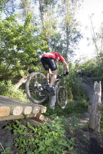 A mountain biker in a red shirt jumps over a wooden ramp in a lush, green trail, surrounded by trees and foliage. Another biker can be seen in the background, navigating the rough terrain. Markham Park mountain bike trail.