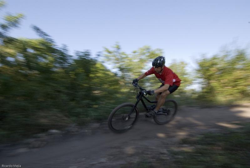 A cyclist wearing a red shirt and helmet is racing along a dirt trail, surrounded by greenery. The image captures the motion blur of the bike and the rider, emphasizing speed and movement in an outdoor setting. Markham Park mountain bike trail.