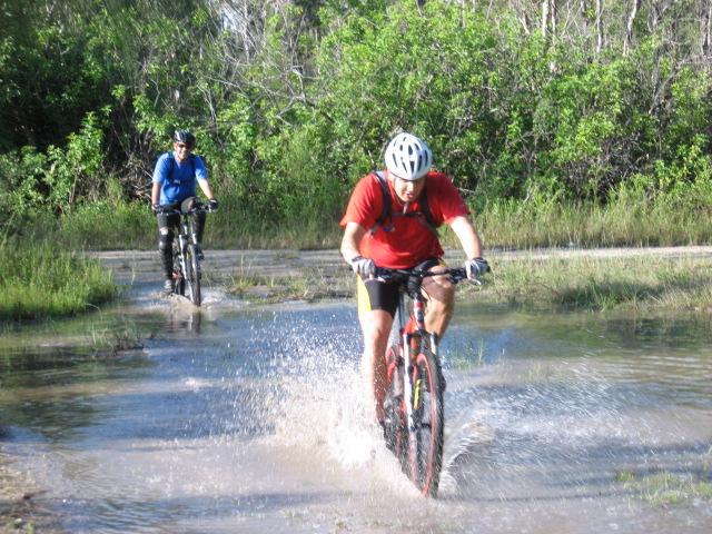 Two mountain bikers riding through a puddle on a dirt path surrounded by greenery. One rider in a red shirt and helmet is splashing through the water, while another rider in a blue shirt follows behind, navigating the trail. Markham Park mountain bike trail.