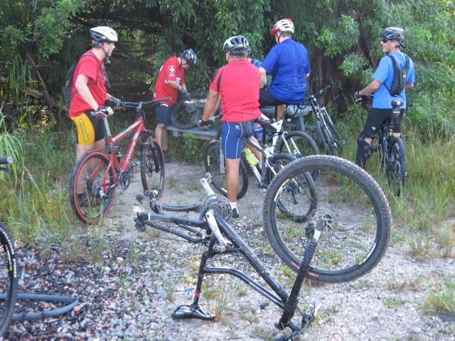 A group of mountain bikers gathered in a natural setting, working on a bike repair. Several riders are wearing helmets and cycling gear, while one bike is turned upside down, revealing its frame and wheels. Surrounding greenery and dirt trail suggest an outdoor adventure. Markham Park mountain bike trail.