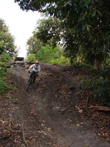 A mountain biker navigating a dirt trail on a slope, surrounded by lush greenery and trees. The rider is leaning forward as they descend, showcasing an action shot of off-road biking. Markham Park mountain bike trail.