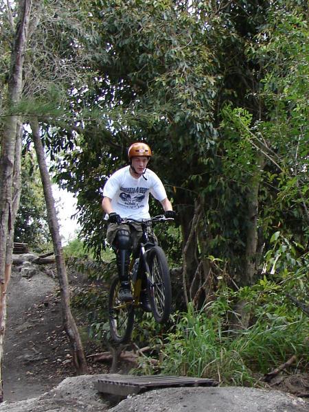 A mountain biker wearing a helmet and a white t-shirt performs a jump on a dirt trail surrounded by trees and foliage. Markham Park mountain bike trail.