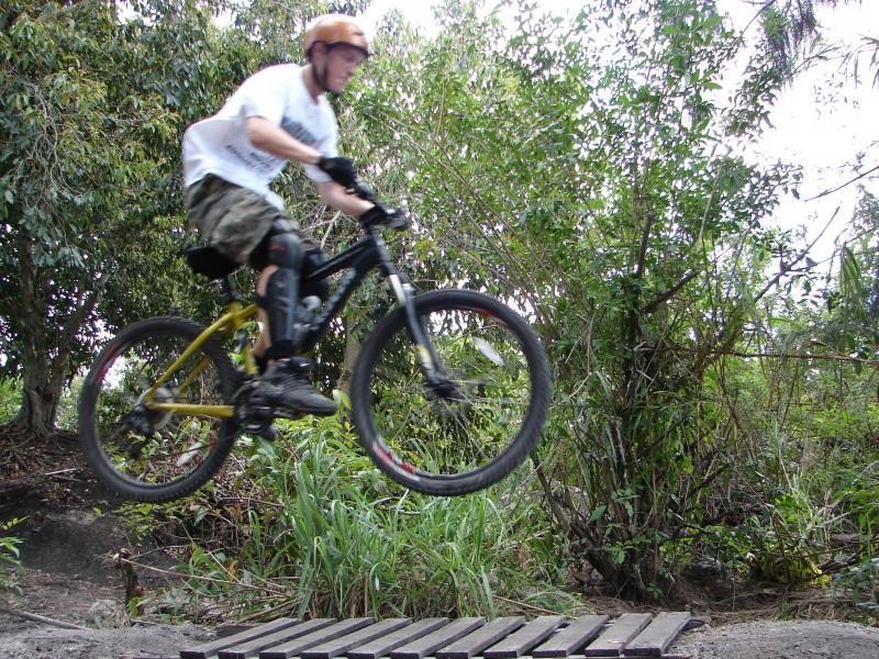A cyclist performing a jump on a mountain bike over a wooden ramp surrounded by greenery. The rider is wearing a helmet, a white t-shirt, and camo shorts, and is captured in mid-air with a dynamic pose. Markham Park mountain bike trail.