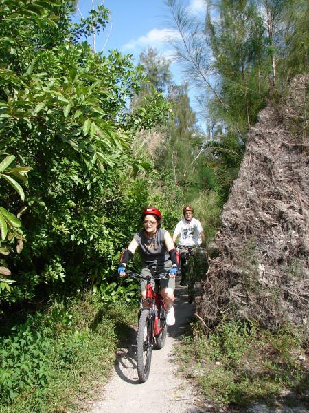Two people riding mountain bikes on a narrow trail surrounded by lush greenery. One rider, wearing a red helmet and sunglasses, is smiling while biking in the foreground, and another cyclist, dressed in casual clothing, follows behind. The scene is set on a sunny day with a blue sky and visible trees. Markham Park mountain bike trail.