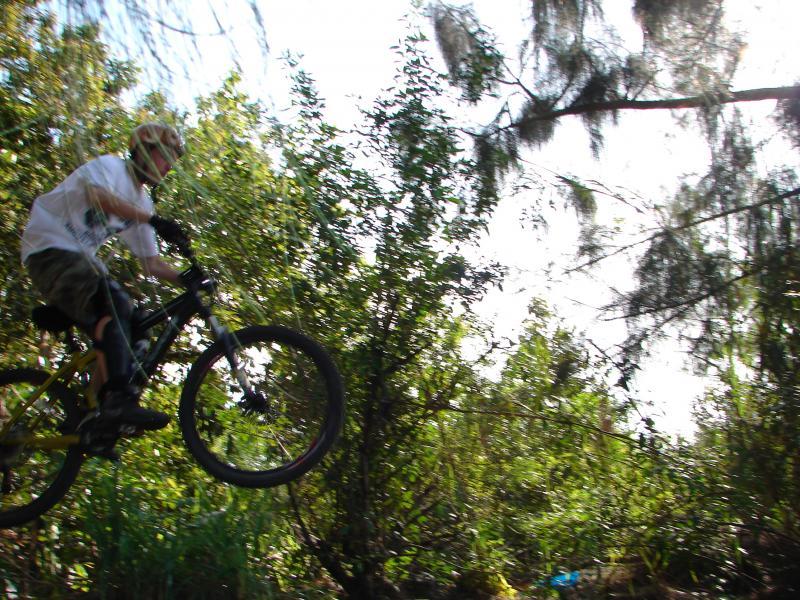 A person riding a mountain bike is captured mid-air while jumping off a natural ramp in a dense, green forest. The rider is wearing a helmet and casual clothing, with sunlight filtering through the trees in the background. Markham Park mountain bike trail.