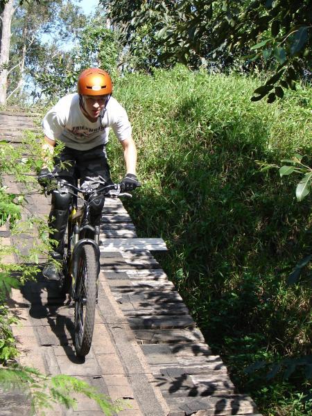 A person wearing an orange helmet and a white t-shirt rides a mountain bike on a narrow wooden path surrounded by greenery and trees. Markham Park mountain bike trail.