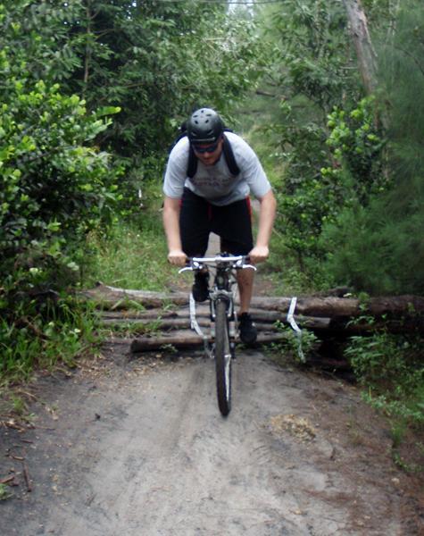 A cyclist wearing a helmet and sunglasses navigates over a log on a dirt trail surrounded by lush greenery. The rider leans forward on the bike, showcasing an action-packed moment in a mountain biking setting. Markham Park mountain bike trail.