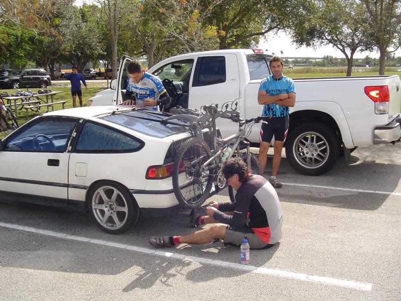 Three cyclists are gathered in a parking area. One cyclist is crouched beside a white car, making adjustments to his bike, while two others stand next to a white pickup truck. The car has a bike rack attached to the back, holding several mountain bikes. Lush trees are visible in the background, indicating a warm, outdoor setting. Markham Park mountain bike trail.