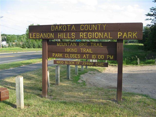 Sign marking the entrance to Lebanon Hills Regional Park in Dakota County, featuring information about mountain biking and hiking trails, park closing time at 10:00 PM, and the address for the 4800 block of Johnny Cake. The surroundings include grass and trees, with a road visible in the background. Lebanon Hills mountain bike trail.